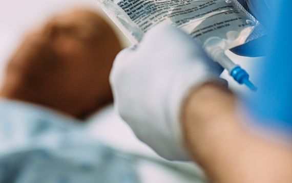 A patient lies in a hospital bed while a nurse looks at an IV bag